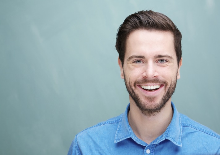 A man with dark hair and a beard smiles at the camera, showcasing his bright smile in a blue denim shirt against a light blue background.